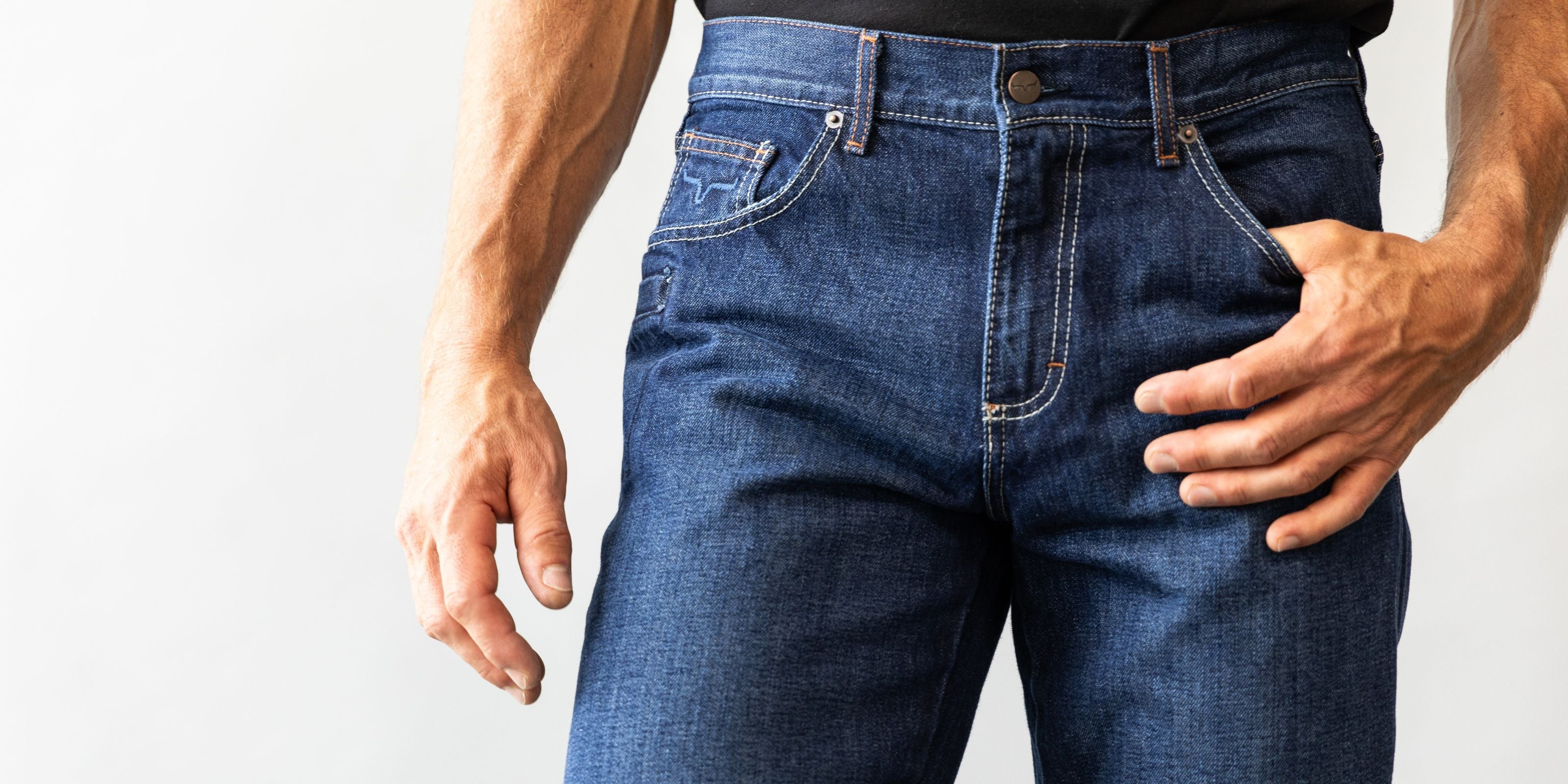 Close-up of a man wearing Kimes Ranch Raw Dillon jeans in unwashed indigo denim with a relaxed fit and mid-low rise. The jeans feature contrast stitching, a horn logo on the front pocket, and a classic Western bootcut style