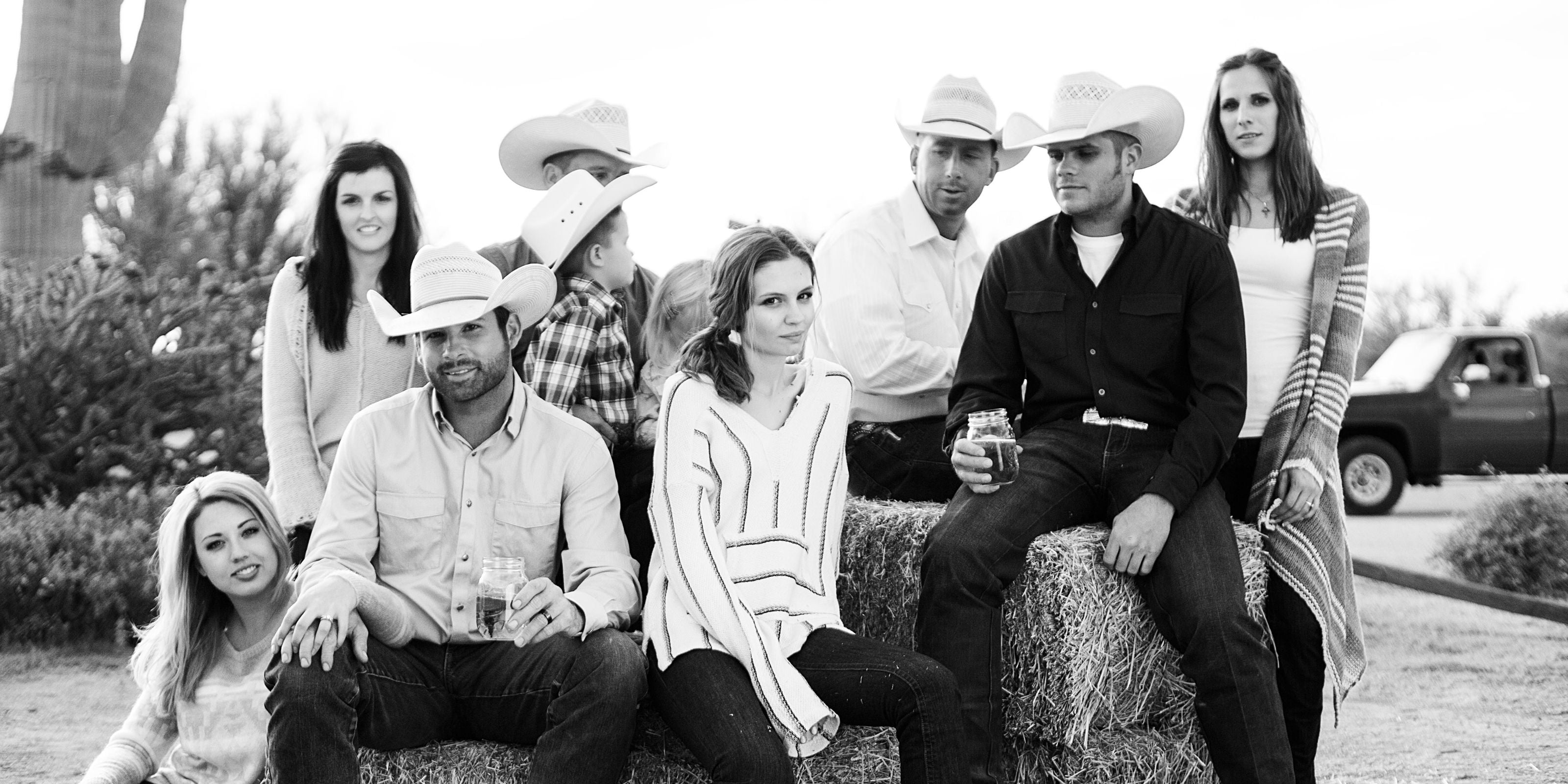 Black and white photo of a group of people wearing Western-style Kimes Ranch clothing, sitting and standing around hay bales outdoors. Some wear cowboy hats and denim, creating a relaxed ranch-inspired atmosphere.