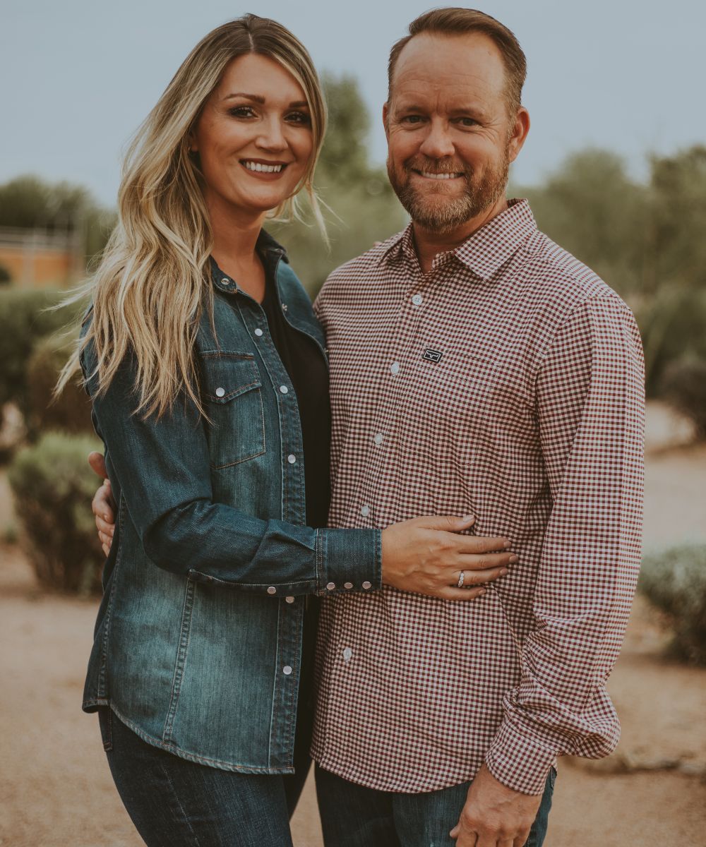 Amanda and Matt Kimes, founders of Kimes Ranch, stand outdoors smiling together. Amanda wears a denim shirt, and Matt wears a red checkered button-down shirt, representing their Western lifestyle brand.