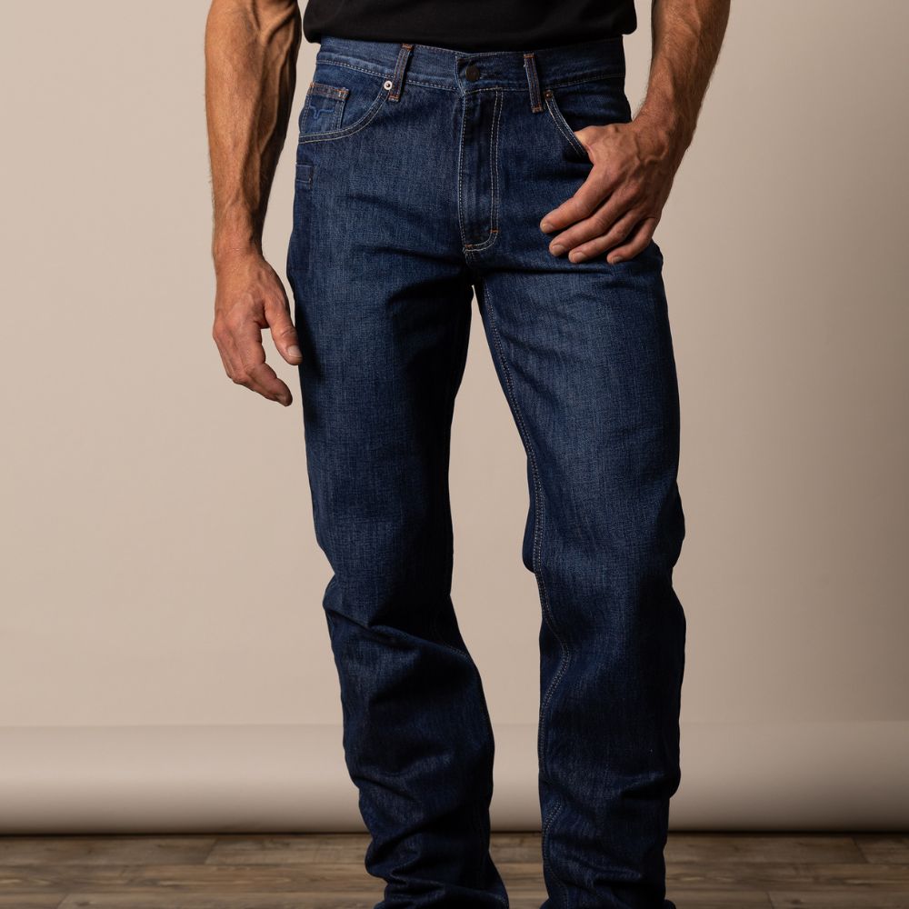 Man wearing Kimes Ranch Raw Dillon jeans in unwashed indigo denim with a relaxed thigh and wide bootcut opening. The jeans feature low-profile blue stitching, an integrated knife pocket, and a rugged Western fit designed for durability and comfort.
