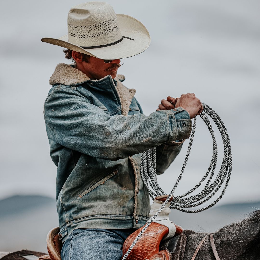 Cowboy wearing the Kimes Ranch Laramie Denim Jacket with a sherpa collar while coiling a rope on horseback. The jacket features a rugged, weathered look that blends durability with classic Western style.