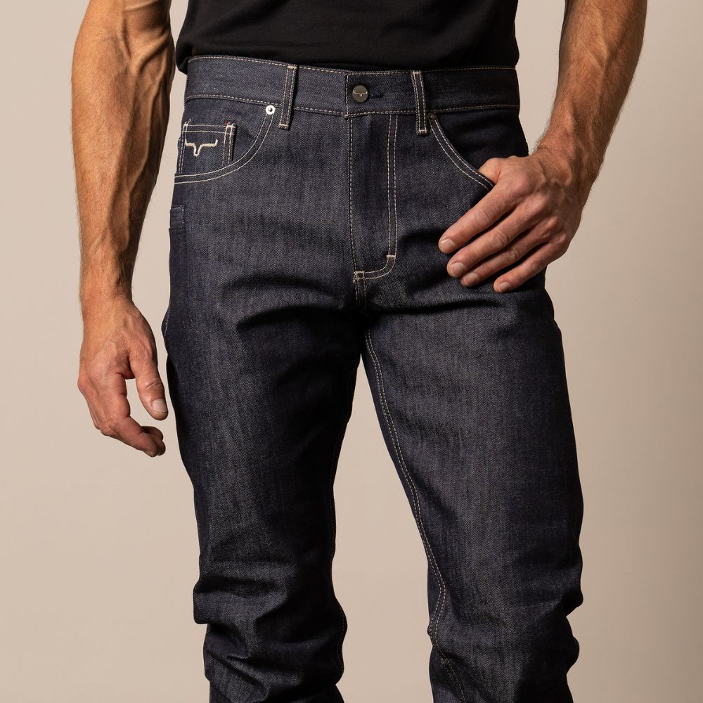 Man wearing Kimes Ranch Raw James jeans in unwashed indigo denim with a mid-low rise and straight bootcut leg. The jeans feature light stitching, an integrated knife pocket, and a classic Western look that molds uniquely with wear.