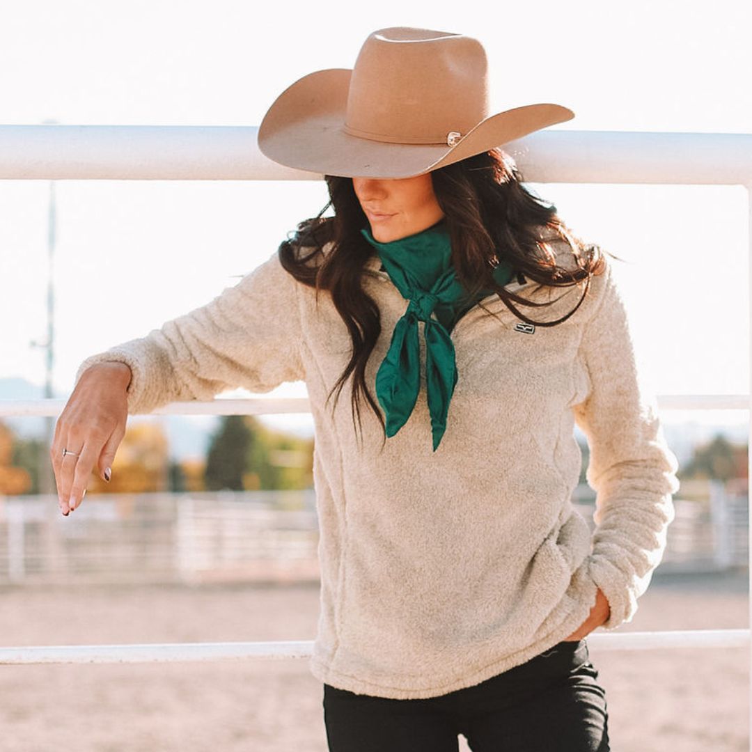 Woman wearing a tan cowboy hat and cream Kimes Ranch fleece pullover, paired with a green scarf. She leans on a white fence in a sunlit outdoor arena, capturing a cozy Western fall look.