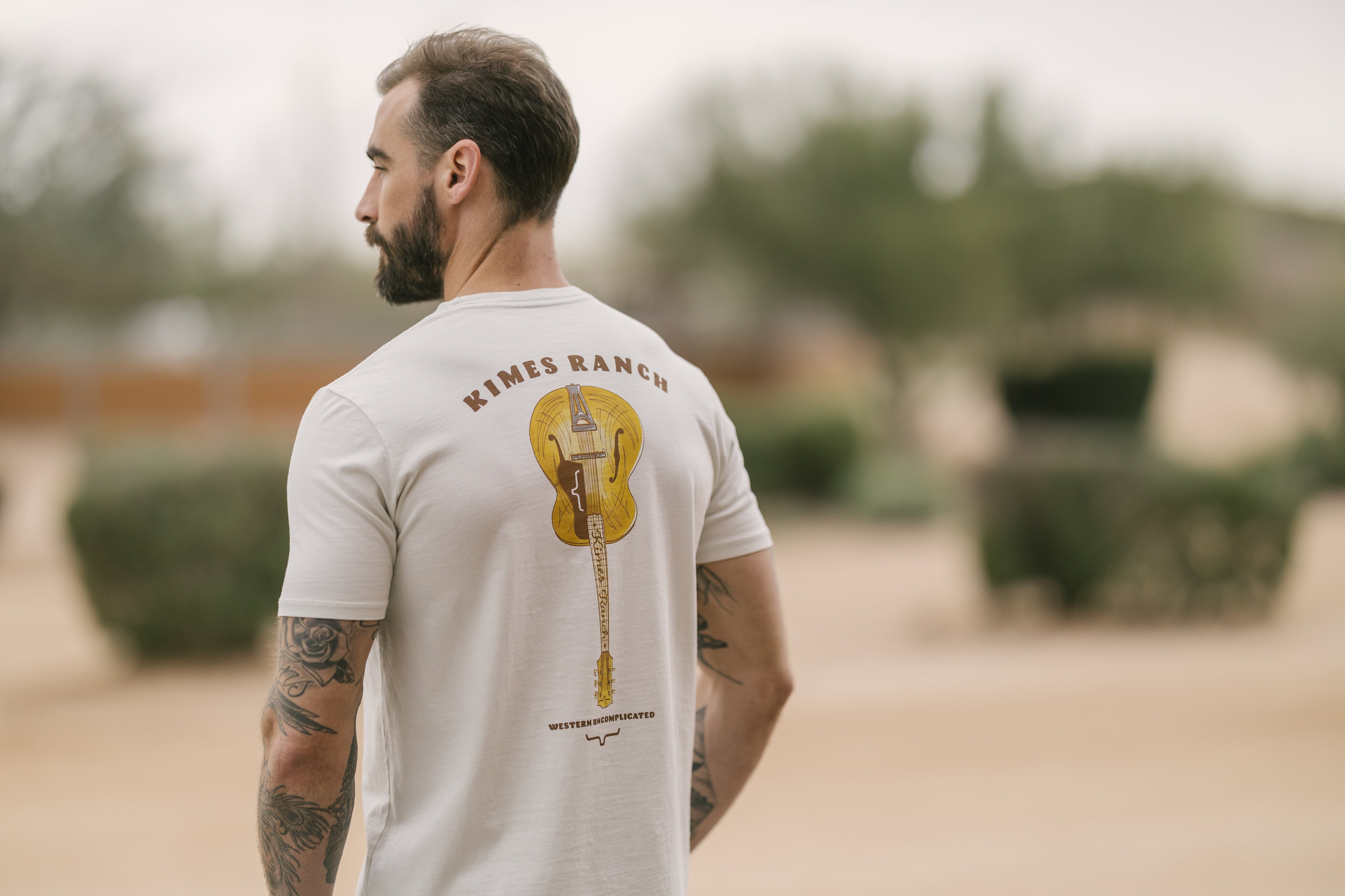 Man wearing a Kimes Ranch western wear graphic tee featuring a guitar design and "Western Uncomplicated" text on the back, shot outdoors on a ranch