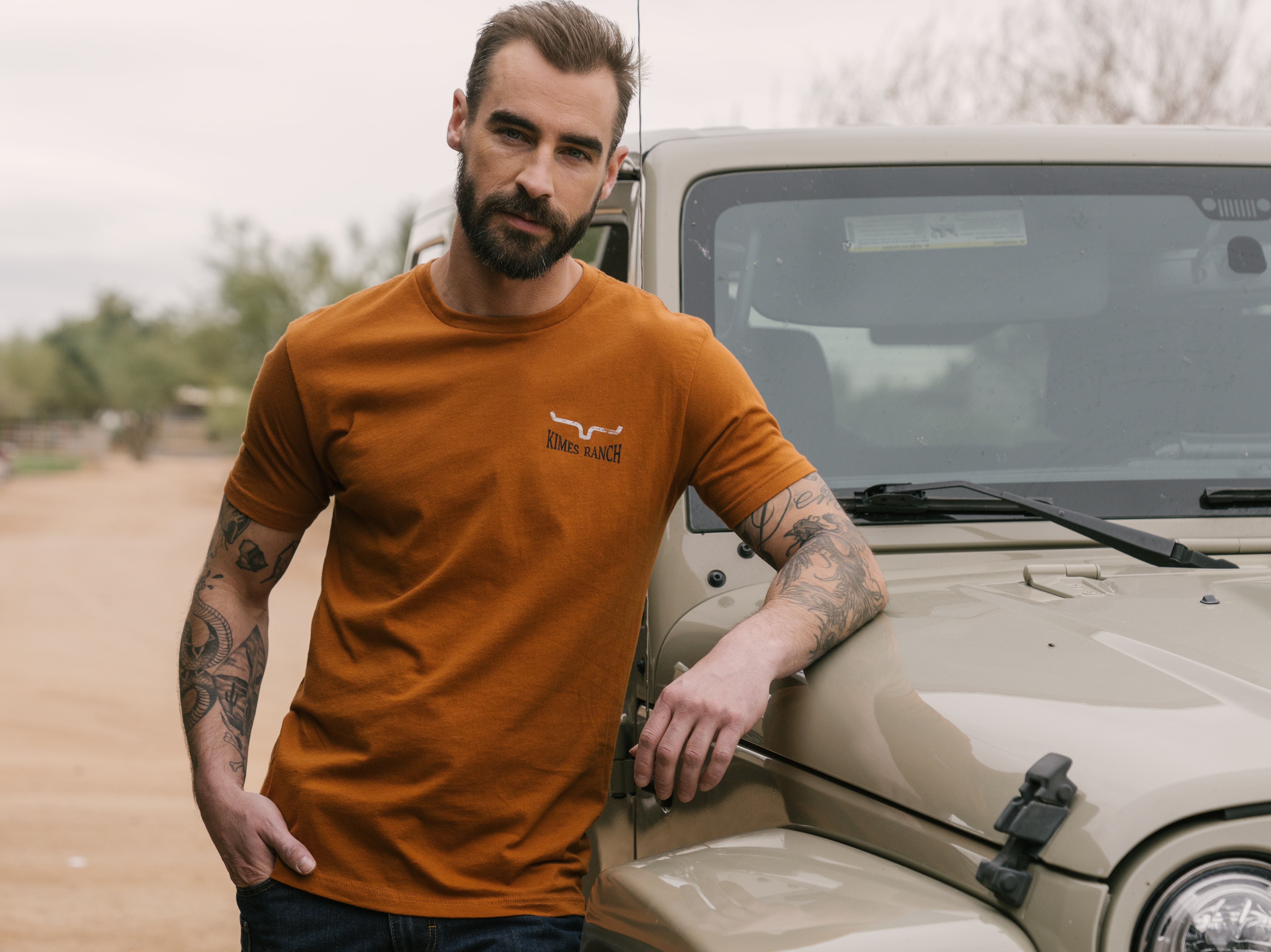 Man wearing a brown Kimes Ranch western wear tee leaning against a Jeep in a ranch setting.